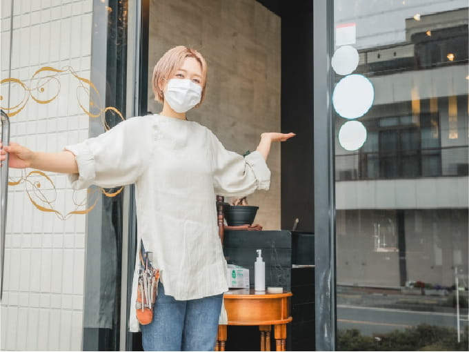 マスクを着用した美容師がサロンの入口で両手を広げて歓迎している様子